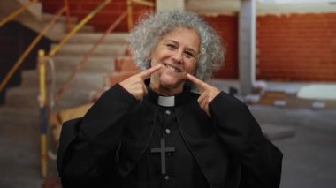 İnşaat alanında gri saç ve çapraz kolyeyle mutlu bir şekilde gülümseyen, siyah rahip kıyafeti giyen, gülümseyen ve bitmemiş dekorun ortasında duran bir kadın..