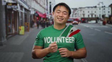 Yeşil gönüllü gömlekli genç adam, Meksika bayrağını elinde tutuyor. Kentsel caddede kültürel çeşitlilik sergiliyor..
