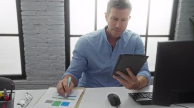 Modern ofiste tablet üzerinde çalışan bir adam, pencerelerden içeri güneş ışığı süzülürken, masa başında evrak ve bilgisayar ekipmanlarıyla uğraşırken dikkatle görevlere odaklanıyor..