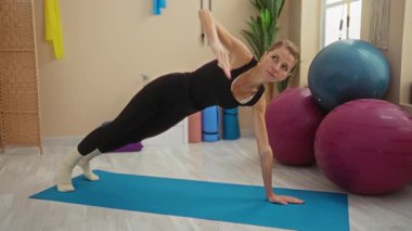 Evde yoga yapan bir kadın spor salonundaki mavi bir paspasın üzerinde egzersiz yapıyor. Egzersiz topları ve etrafındaki canlı ekipmanlarla görülebiliyor..