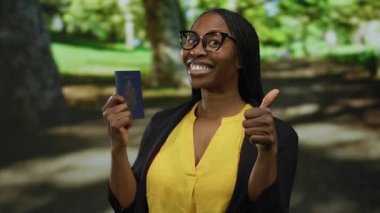 Parkta elinde Kanada pasaportuyla gülümseyen bir kadın seyahat özgürlüğünü ve heyecanı simgeliyor. Gözlük takıyor ve parlak sarı bir üst ve bulanık ağaçlar..