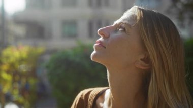 Şehir parkında güneşli şehir manzarasına bakan bir kadın. Sarışın saçlı, meraklı ve düşünceli bir kadın. Huzurlu bir çevrede gençlik ve güzelliği vurguluyor..
