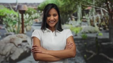 Parkta kaktüs bitkileriyle gülümseyen genç bir kadın, kollarını kavuşturmuş, günlük beyaz giyinmiş, açık havada, mutluluk ve kendine güvenini gösteriyor..