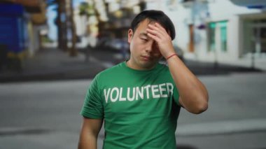 Yeşil gönüllü tişörtü giyen genç adam, arka planda bulanık binalar olan hareketli bir şehir caddesinde dikilirken düşünceli görünüyor..