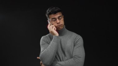 Gözlüklü genç adam, koyu bir arka plan karşısında derin bir ifadeyle akıllı telefonda konuşuyor, gri balıkçı yaka giyiyor ve düşünceli vücut dilini gösteriyor.