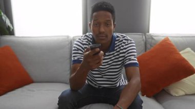 Çizgili gömlekli genç adam oturma odasında kanepede oturur. Gülümser ve uzaktan kumandayı gösterir. Rahat ev atmosferini turuncu yastıklarla taşır..