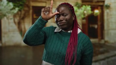 Örgü örgülü Afro-Amerikalı kadın, yeşil kazak giyen ve kameraya gülümseyen, şehir dışında hayat dolu bir sahne yaratan ezik bir sembol gösteriyor..