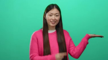 Pembe kazaklı kadın, parlak yeşil bir arka planda el kol hareketi yapıyor, etkileyici bir poz sergiliyor ve çekici bir gülümseme sergiliyor, neşeli bir ortamda kendine güveni ve coşkuyu somutlaştırıyor..