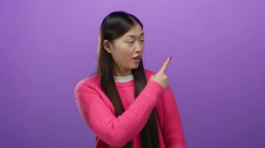 Pembe kazak giyen kadın mor arkaplanı işaret ediyor, canlı renk kontrastını vurguluyor ve izole ortama karşı etkileyici hareketleriyle dikkat çekiyor..