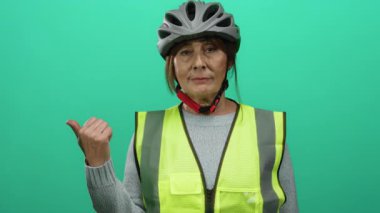 Yeşil arka plana karşı miğfer ve güvenlik yeleği giyerken başparmağıyla işaret eden kadın, güvenli açık hava faaliyetlerine güven ve güvenin bir göstergesi..