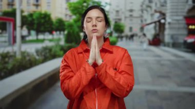 Elleri kelepçeli turuncu elbiseli bir kadın şehir sokaklarında dua ediyor, gözleri kapalı ve yüzleri barışçıl, kentsel binalar ve yeşilliklerle çevrili..