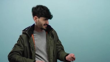 Mavi bir arka plana hayali bir ip çeken genç bir adam, yeşil bir ceket giyiyor ve neşeli görünüyor, yüzünde odaklanmış bir ifadeyle çevreden soyutlanmış..