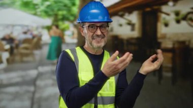 Şehir parkında mavi miğfer ve sarı yelek giyen bir adam, elleriyle el kol hareketi yapıyor, açık şehir ortamında kendine güven ve bağlılık yayıyor..