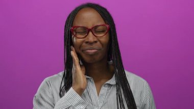 Dişi ağrıyan bir kadın kıvırcık saçlı ve gözlüklü canlı pembe bir arka plana karşı duruyor. Acı ve rahatsızlık ifade ediyor..