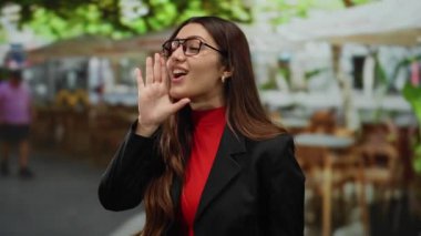 Güneşli bir günde masaların ve ağaçların bulanık arka planına sahip bir restoranın terasında kırmızı ve siyah gözlüklü bir kadın konuşuyor..