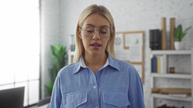 Gözlüklü ve mavi tişörtlü bir kadın, kendine güvenerek parlak bir ofiste duruyor. Çiçekler ve raflarla, profesyonel ve sakin bir tavır sergiliyor..