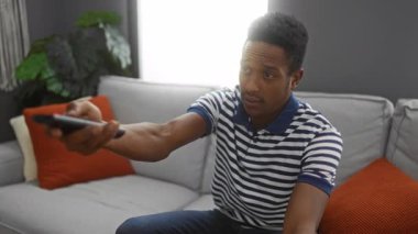 Young man relaxing on a sofa in a cozy living room, holding a remote control, surrounded by cushions and plants, enjoying leisure time indoors.