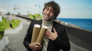 Deniz kenarında gülümseyen genç bir adam elinde iki kağıt tüp ile, arka planda okyanus olan açık deniz ortamında kendini rahat ve mutlu hissediyordu..