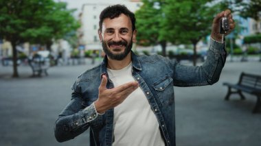 Sokakta elinde araba anahtarı olan İspanyol bir adam yeşil ağaçların altında kot ceket ve beyaz gömlek giyen neşeli bir gülümsemeyle işaret ediyor..