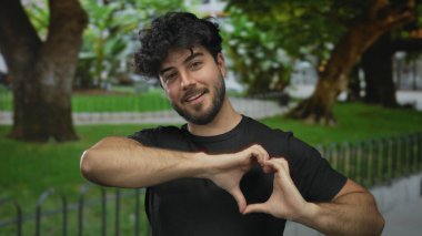 Sakallı genç adam, açık yeşil bir parkta gülümserken kalp hareketi yapıyor, sevgi ve mutluluk taşıyor..