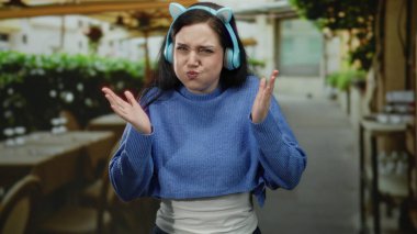 Esmer saçlı, kulaklık takan bir kadın, şehir manzarasıyla çevrili bir restoran terasında sürpriz yaparak, canlı ve modern bir sokak atmosferi yaratıyor..