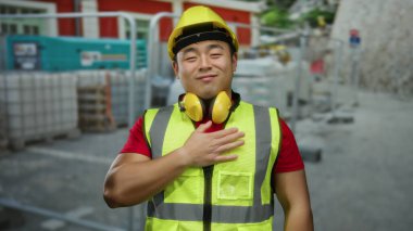 Güvenlik donanımlı genç adam, sarı bir kask ve yelek giymiş, Asyalı kökenini gösteren, sıcaklık ve memnuniyet saçan bir inşaat alanında şükranlarını sunuyor..
