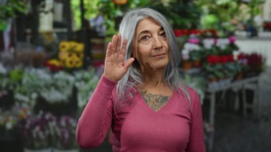 Gri saçlı bir kadın arka planda canlı çiçeklerle açık bir çiçekçide el sallıyor ve dostça bir jest yapıyor..
