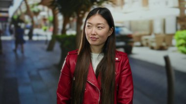 Kırmızı ceketli kadın, şehir arka planında bulanık bir şekilde gülümsüyor. Canlı, rahat ve gençlik tarzını ön plana çıkarıyor..