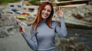 Deniz kenarında elinde İsveç kronuyla gülümseyen bir kadın rahatlatıcı ve mutlu bir ifadeyle iyi bir jest yapıyor. Deniz kenarında başarı ve keyif göstergesi..