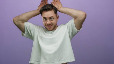 Sakallı genç bir adam, mor bir arka planda, elleri başının üstünde, eğlenceli, kaprisli bir ifade ve neşe sahnesi yaratıyor.