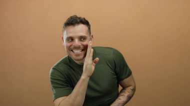 Genç adam açık kahverengi bir arka plan önünde şakacı bir ifadeyle fısıldıyor, yeşil bir gömlek giyiyor ve kollarında dövmeler gösteriyor, gizlilik ve entrika anlatıyor..