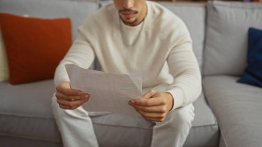 Genç İspanyol bir adam rahat bir oturma odasında oturmuş bir belge okuyor ve evde günlük beyaz giysiler giyiyor..