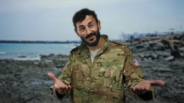 Askeri üniformalı İspanyol bir adam kayalık sahilde deniz zeminiyle duruyor, kendinden emin ve anlamlı görünüyor, askeri varlığı sakin kıyı manzarasıyla harmanlıyor..