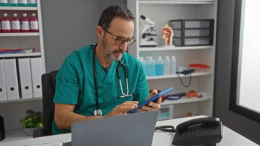 Hastane odasında laptopun ve telefonun yanında tablet kullanan, yeşil önlük giyen, klinik ortamda olgun bir profesyoneli gösteren İspanyol bir adam..