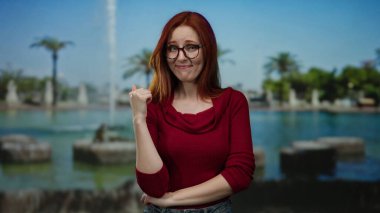 Parkta gözlüklü ve kızıl saçlı bir kadın, kırmızı bir süveter giyiyor, neşeli görünüyor ve başparmağıyla bir çeşme ve yeşillik zeminine işaret ediyor..