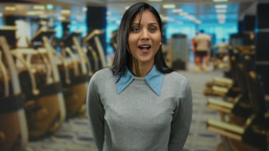 Spor salonunda gülümseyen genç bir kadın gri süveter ve mavi yakalı, arka planda insanlar olan egzersiz ekipmanlarıyla çevrili, canlı bir atmosfer yaratıyor..