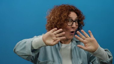 İspanyol, orta yaşlı, kıvırcık saçlı bir kadın kameraya, izole edilmiş mavi bir arkaplana, gözlüklü ve pozitif enerji gösteren günlük gömlekli bir kadına coşkuyla el sallıyor..