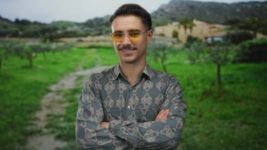 Genç, Latin asıllı bir adam, gür bir park manzarasında güvenle duruyor, kollarını kavuşturup gülümsüyor, şık gözlükler takıyor ve rahat bir açık hava havası yayıyor..