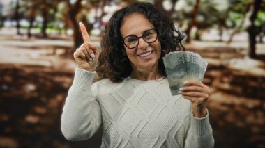 Orta yaşlı bir kadın elinde Endonezya rupiası banknotları tutarken şehir caddesinde durup finansal bir fırsat ya da fikri işaret ediyor..