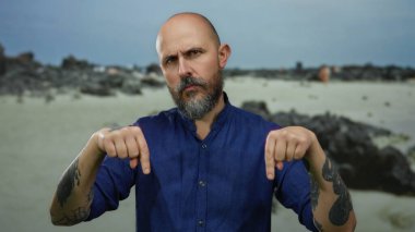 Sakallı bir adam bulutlu bir gökyüzünün altında mavi bir gömlek giyerken kayalık bir sahilde güven içinde el kol hareketi yaparak engebeli bir çevreyi gösteriyor..