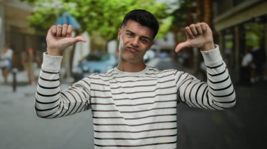 Çizgili gömlekli genç adam şehir caddesinde bulanık ağaçlar ve insanlarla başparmak kaldırıyor. Açık hava ortamını tasvip etmiyor veya hayal kırıklığına uğratmıyor..