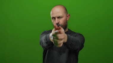 Kel, beyaz, sakallı, yeşil arka planda parmak silahlarıyla poz veren kararlı ve odaklanmış bir adam.