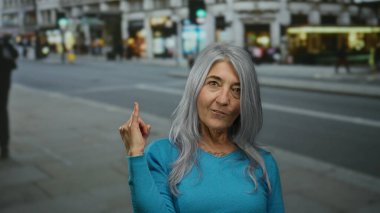 Gri saçlı yaşlı kadın hareketli bir sokakta taş jest yapıyor, mavi bir kazak giyiyor, gençliği ve özgürlüğü sembolize ediyor.