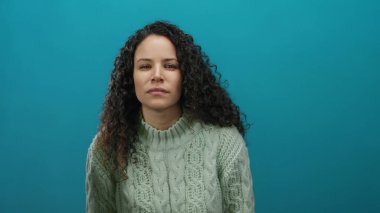 Kazaklı, kıvırcık saçlı bir kadın izole edilmiş mavi duvara karşı duruyor. Düşünceli ve anlamlı görünüyor. Basit ama canlı bir ortamda bir dizi duygu taşıyor..