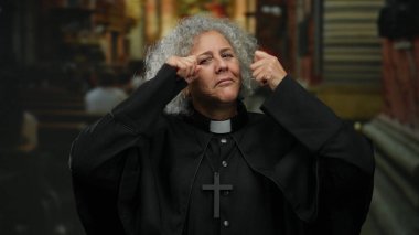 Kır saçlı yaşlı bir kadın, kilise ortamında duygusal jestler yapıyor, papaz kıyafeti ve haç giyiyor, evde maneviyat ve yansıma hissi taşıyor..