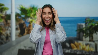 Genç sarışın kadın, neşeli bir şekilde, canlı mavi okyanus arkaplanı ve görünen palmiye ağaçları ile güneşli bir sahil geçidinde surat yapıyor..