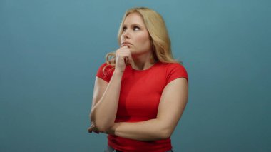 Düşünceli bir ifadeyle düşünen, mavi arka planda kırmızı tişört giyen bir kadın derin düşünme ve karar verme anını resmediyor..