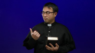 Elinde incili tutan rahip mavi arka planda düşünceli bir ifade sergiliyor.