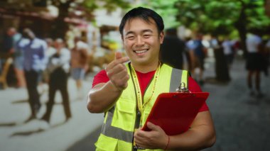 Güvenlik yeleği giyen genç adam, parmaklarıyla kalp hareketi yapıyor, kalabalık bir şehir arka planında gülümsüyor..