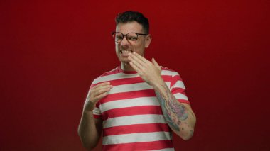 Dövmeli, gözlüklü, çizgili tişörtlü, kırmızı arka planda duygularını ifade eden bir adam..